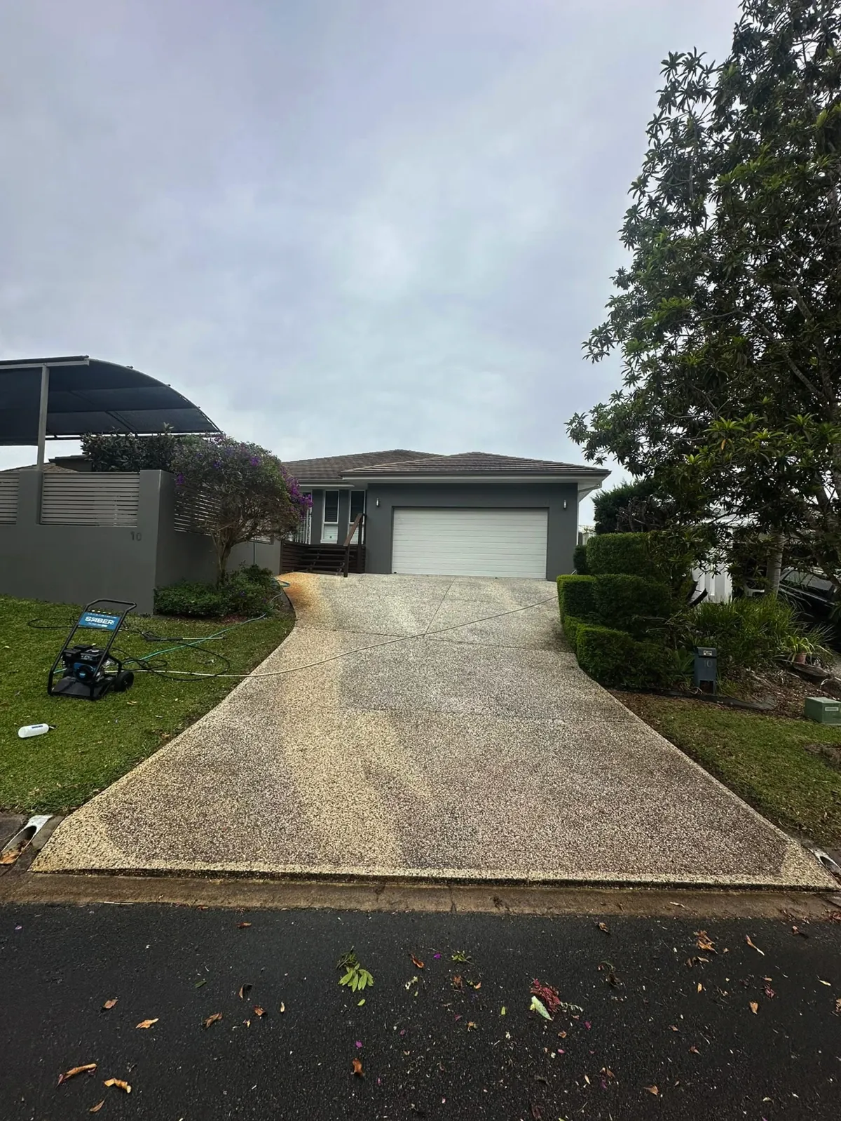 An exposed aggregate driveway before cleaning, grey and stained, with the pressure washer set up on the lawn